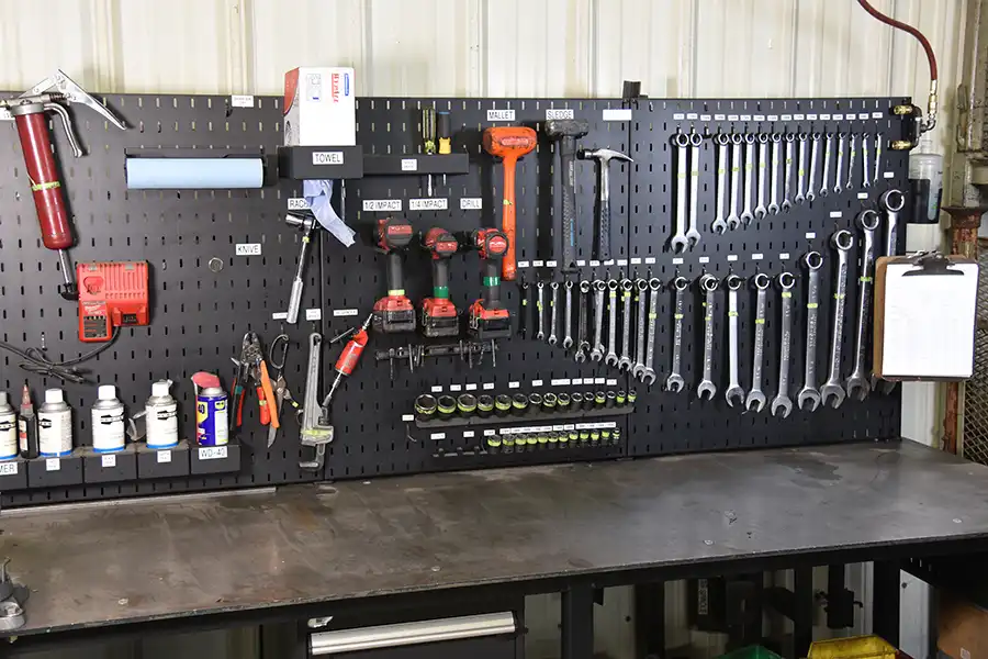 A black pegboard above a workbench displays organized tools, demonstrating 5 Key Features of a 5S Workstation. Wrenches, drills, screwdrivers, and labeled bottles are neatly arranged, with a clipboard on the right and the workbench surface mostly clear.